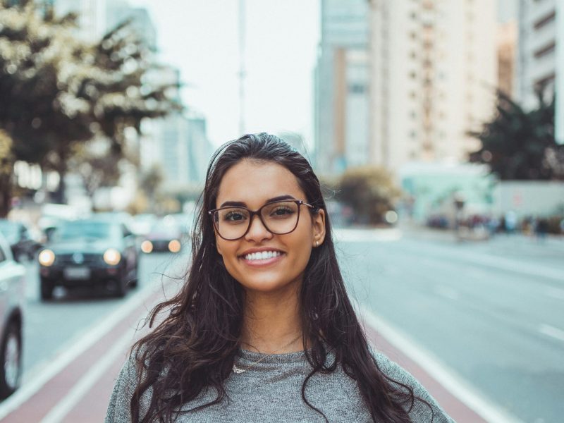 A young woman with glasses smiling on a city street, embracing urban lifestyle.