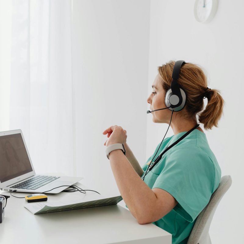 A female doctor using a laptop for an online consultation, wearing a headset in a bright office.